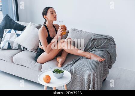 Young woman with slim body shape in sportswear sits on sofa and eats healthy diet food indoors at home Stock Photo