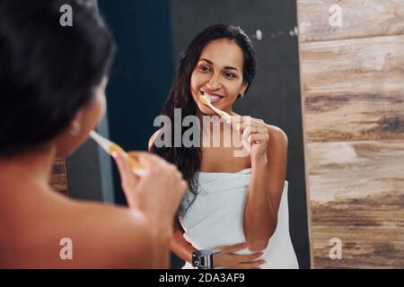 Beautiful young brunette standing in bathroom near the mirror and brushing her teeth. Stock Photo