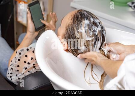 woman in a beauty salon. hair care. hairdresser applies mask to client's hair. female looking at her mobile phone Stock Photo