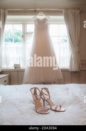 Bride's wedding dress hanging at a bedroom window with shoes on the bed in the foreground. Stock Photo
