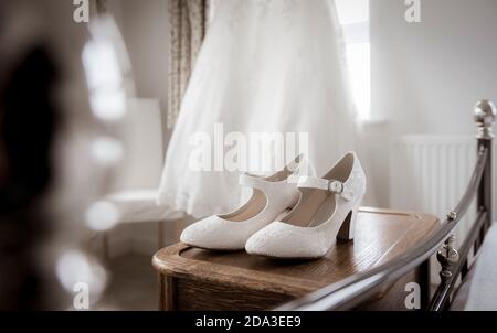 Bride's wedding dress hanging at a bedroom window with shoes in the foreground. Stock Photo