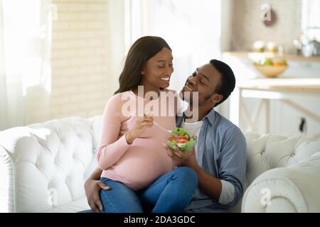 Balanced diet in pregnancy concept. Handsome black man giving his lovely expectant wife yummy vegetable salad indoors Stock Photo