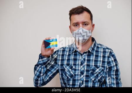 Man in checkered shirt show The Bahamas flag card in hand, wear protect mask isolated on white background. American countries Coronavirus concept. Stock Photo