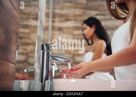 Beautiful young woman standing in bathroom near the mirror and washing hands. Friends behind Stock Photo