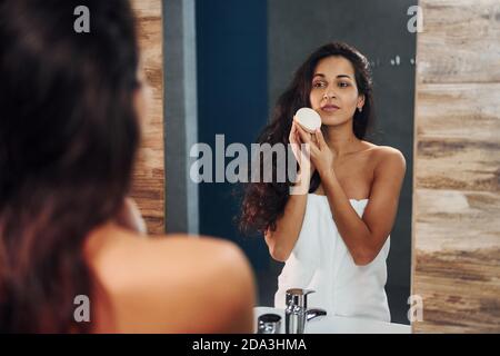 Beautiful brunette standing in bathroom near the mirror and cleaning her face Stock Photo