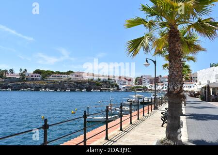 promenade in a village on the island of Menorca in summer sunshine, with boats and palm trees lining the road Stock Photo