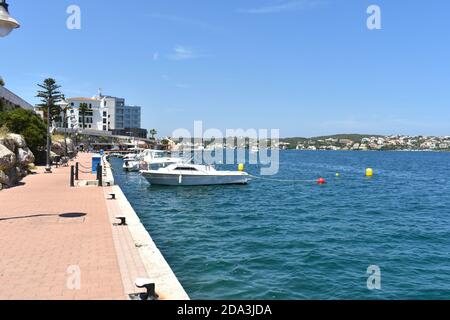 promenade in a village on the island of Menorca in summer sunshine, with boats and palm trees lining the road Stock Photo