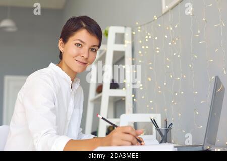 Smiling woman sitting with laptop and making notes during video call or online lesson at home Stock Photo