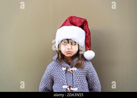 Sad little girl wearing Santa Claus hat against a green wall background outdoors in christmas time - Little girl sulking for unwanted xmas gift - Unha Stock Photo