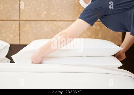 Cropped of maid in mask adjusting white pillows on the bed in hotel room. Hotel service concept Stock Photo