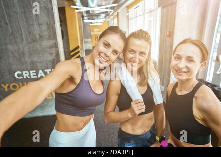 Working out with friends. Group of three young sportive women taking selfie on smartphone, looking at camera and smiling while standing at gym. Sport, wellness and healthy lifestyle Stock Photo