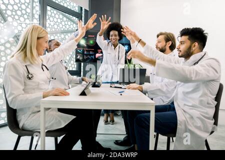 Group of motivated international doctors or scientists, sitting around the table in conference room at modern clinic, giving a high fives gesture with Stock Photo