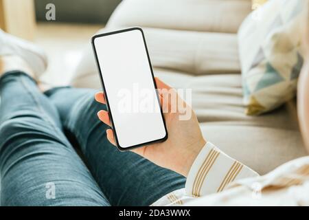 Mockup of a phone held by a woman's hand who is sitting on the couch. White screen Stock Photo
