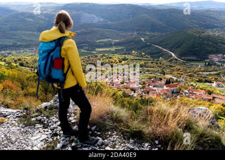 Woman wearing backpack standing on a cliff with a view of Crni Kal village in Slovenia Stock Photo