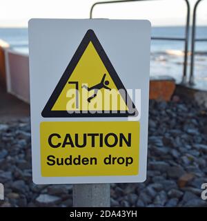 Yellow, black and white sign Caution Sudden drop with warning triangle and icon of person falling over edge. Blurred harbour background. Stock Photo