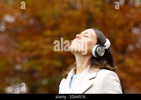 Middle age relaxed woman breathing deeply fresh air wearing headphones in a forest in autumn