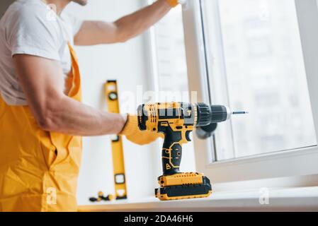 Handyman in yellow uniform installs new window by using automatic screwdriver. House renovation conception Stock Photo