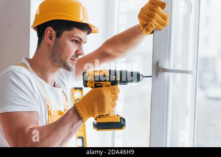 Handyman in yellow uniform installs new window by using automatic screwdriver. House renovation conception Stock Photo