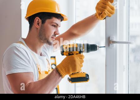 Handyman in yellow uniform installs new window by using automatic screwdriver. House renovation conception Stock Photo