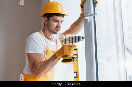 Handyman in yellow uniform installs new window by using automatic screwdriver. House renovation conception Stock Photo