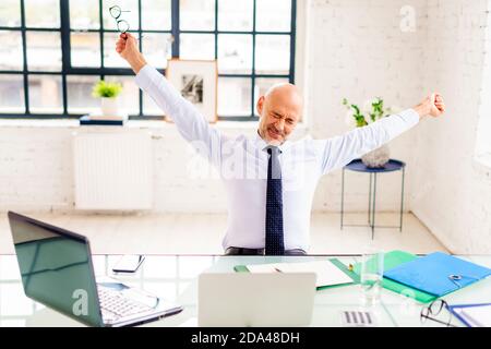 Portrait shot of frustrated businessman with raising hands sitting at office desk behind his laptop while working online. Stock Photo