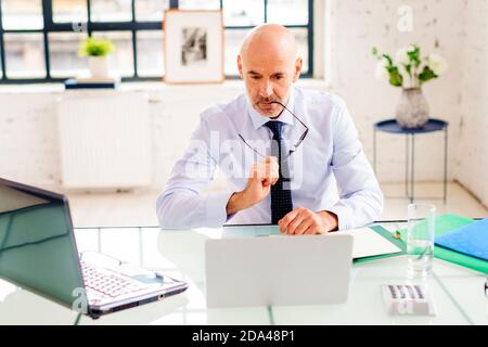 Portrait shot of unhappy senior businessman looking thoughtfully while sitting at desk and working on laptop at the office. Stock Photo
