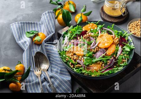 Fresh homemade tangerine and beet salad with arugula, walnuts and pine nuts on grey concrete background with dressing and ingredients. Healthy vegan f Stock Photo