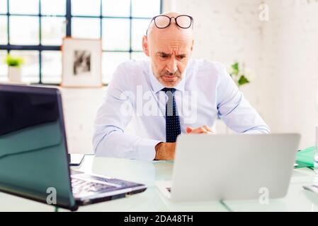 Portrait shot of unhappy senior businessman sitting at desk and using laptop while working at the office. Stock Photo