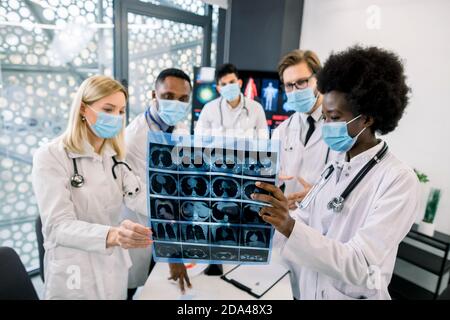 Team of multiethnic doctors or scientists, wearing protective medical face masks, looking at the CT scan of the patient and discussing the results of Stock Photo
