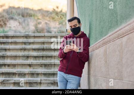 Young man with a mask writes on his phone leaning against a wall Stock Photo