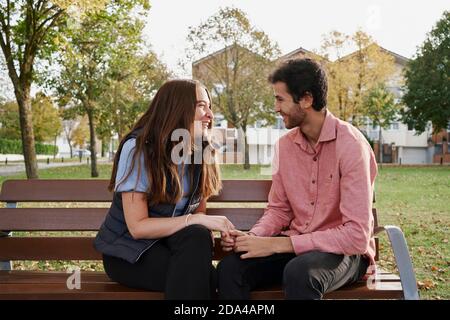 Young couple smiling and staring at each other sitting on the bench in a park. Romantic concept Stock Photo