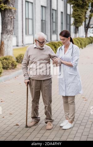 brunette social worker helping elderly man with walking stick while strolling together Stock Photo