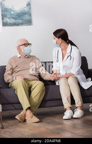 nurse with stethoscope holding hand of elderly man on sofa at home Stock Photo