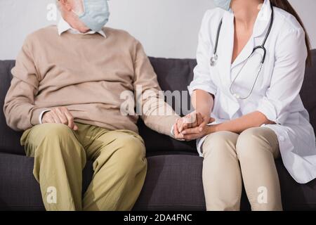 partial view of social worker in medical mask holding hands of aged man at home Stock Photo