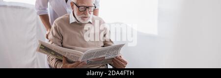 aged man in eyeglasses reading newspaper near social worker at home, banner Stock Photo