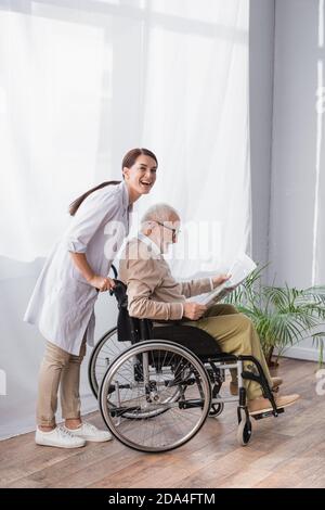 geriatric nurse smiling at camera near disabled elderly man reading newspaper in wheelchair Stock Photo