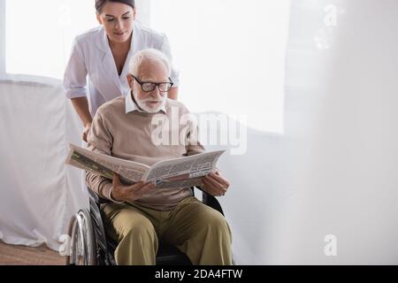 geriatric nurse near elderly handicapped man reading newspaper in wheelchair Stock Photo