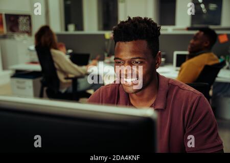 African male business man smiling while typing on desktop sitting in office with colleagues  Stock Photo