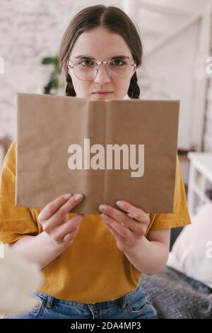Young beautiful woman student in orange T-shirt and glasses reads book sitting at the table. Preparing for exams, studying at home on self-isolation Stock Photo
