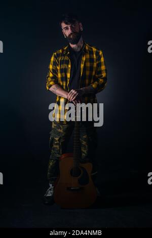 Tattooed man with a beard wearing a tartan yellow black shirt is standing and posing with his guitar in his hand Stock Photo