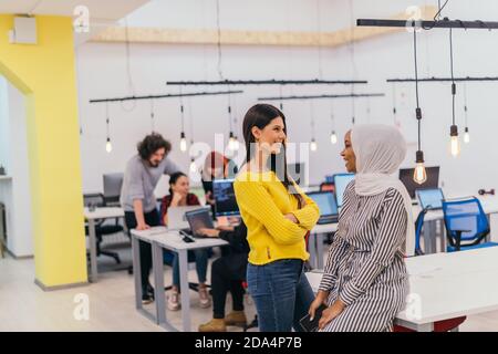 Portrait of two African American businesswomen talking to each other while standing in a modern business office with their colleagues, coworkers in th Stock Photo