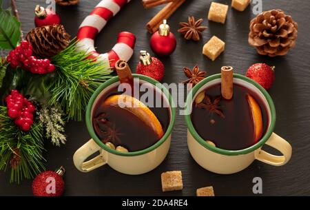 Mulled wine, winter warming alcohol drink, two vintage mugs, ingredients orange and spices and Christmas decoration on black stone, closeup view. Stock Photo
