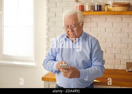 Serious senior man in glasses standing in the kitchen and using modern mobile phone Stock Photo