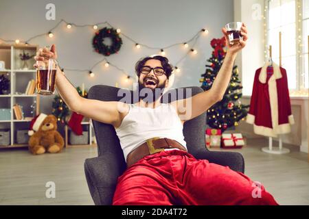 Happy young man in tank top and Santa costume relaxing in armchair at home and drinking whiskey Stock Photo