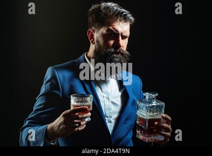 Bartender leather apron holding whisky cocktail in glass. Hipster with beard and mustache in suit drinks alcohol after working day. Stock Photo