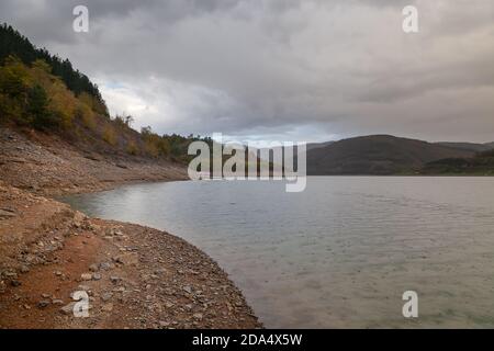 Raindrops falling on the surface of beautiful, clean, fresh water Zavoj lake, distant boat, red rocky foreground and murky sky Stock Photo