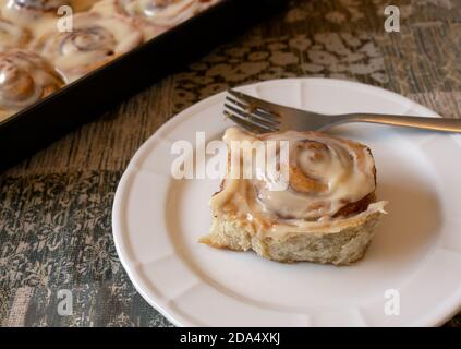 Cinnamon rolls with cream cheese, over white plate with fork and baking pan in the background Stock Photo