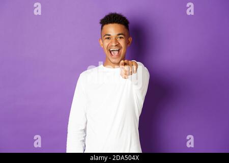 Image of handsome young african-american man in white sweatshirt, pointing finger at camera and smiling, choosing you, standing over purple background Stock Photo