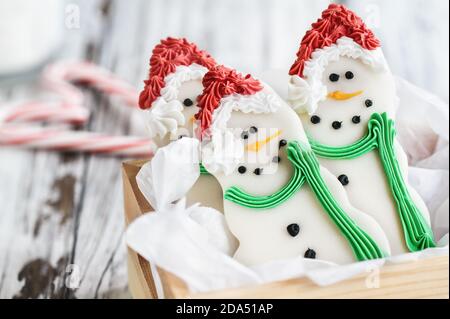 Iced Christmas Snowman cookies or biscuits with carrot nose, santa hat, and scarf packaged in a wood box with candy cane heart behind them. Selective Stock Photo