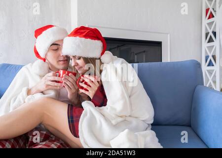 Young Caucasian couple drinking hot tea or cocoa from red cups while sitting at home on sofa wearing santa hats and pajamas wrapped in white blanket Stock Photo